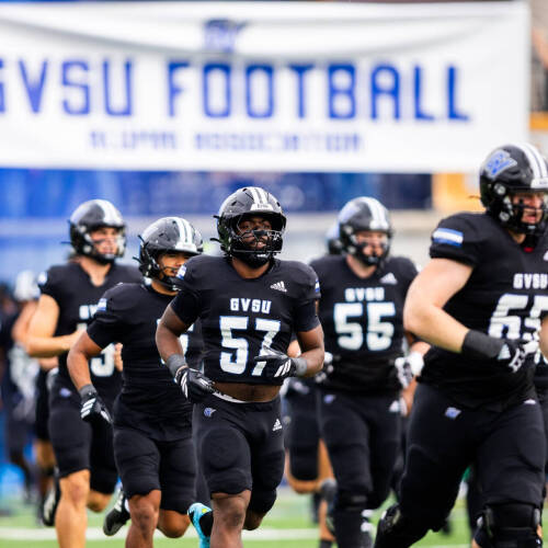 Laker football team runs under GVSU Football banner wearing all black with black helmets